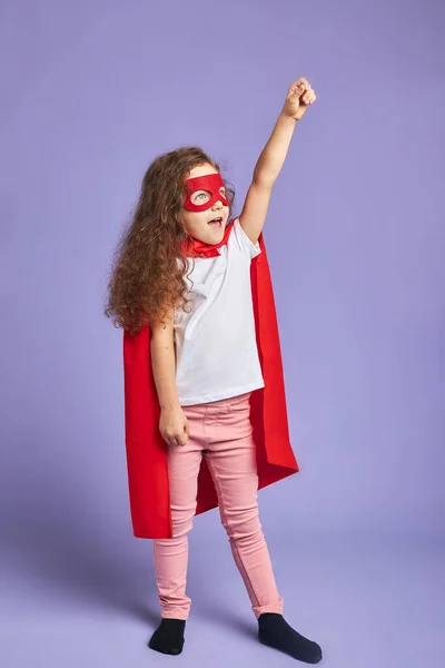 Alegre niña pequeña con máscara roja y capa aislado fondo púrpura — Foto de Stock