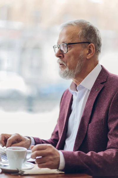 Allvarlig skäggig man i röd elegant kostym sitter vid bordet och surfar på nätet — Stockfoto