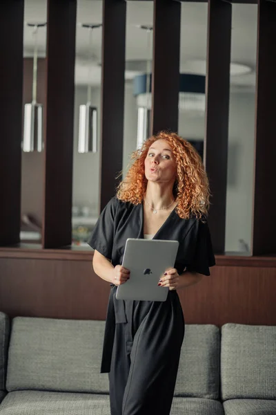 Mulher em macacão preto elegante com cabelo de gengibre encaracolado posando com laptop — Fotografia de Stock