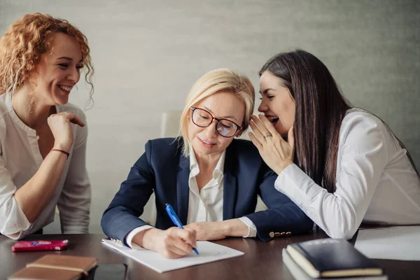 Mujer de negocios escribiendo un documento y dos compañeras de trabajo chismeando en sus oídos — Foto de Stock