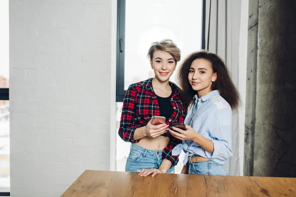 Dos atractiva rubia y morena están utilizando teléfonos móviles en la sala de estar — Foto de Stock