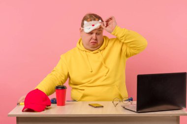 Uykucu adam dizüstü bilgisayarın önünde elinde kahveyle masada oturuyor.