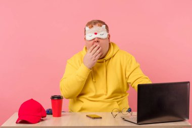 Uykucu adam dizüstü bilgisayarın önünde elinde kahveyle masada oturuyor.