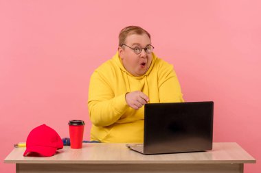 Şişman vücutlu utangaç bir adam ağzını açar ve dizüstü bilgisayarını gösterir..