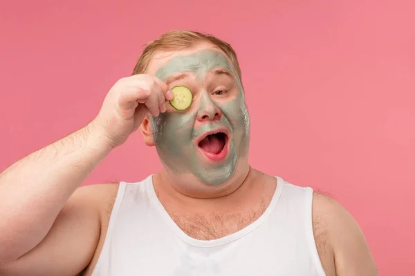 Homem feliz com máscara de barro facial para cuidados com a pele se divertindo com fatias de pepino — Fotografia de Stock