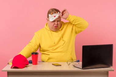 Uykucu adam dizüstü bilgisayarın önünde elinde kahveyle masada oturuyor.