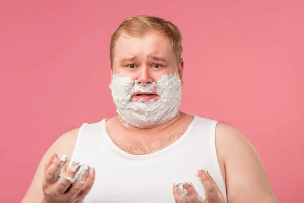 Sad man with gel foam on cheeks, applying foam on skin before shaving morning.