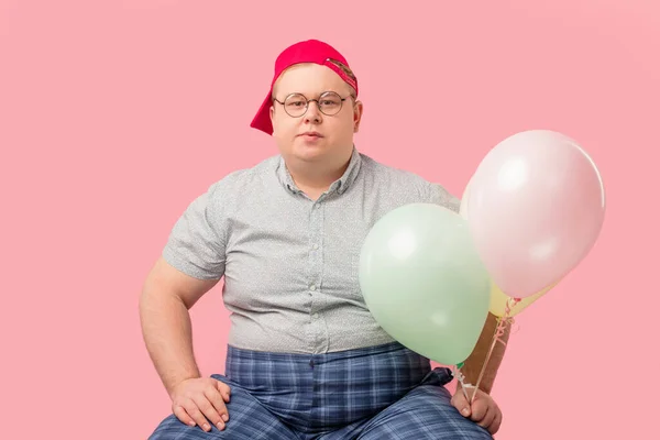 Geschwollener erwachsener Mann mit roter Baseballkappe und karierter Hose posiert mit Luftballons, — Stockfoto