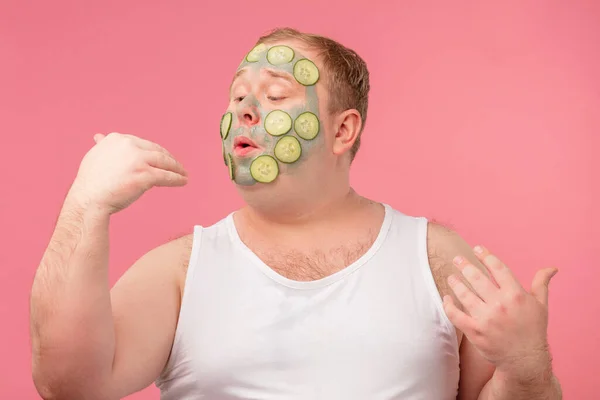 Hombre feliz con máscara de arcilla facial para el cuidado de la piel que se divierte con rodajas de pepino — Foto de Stock