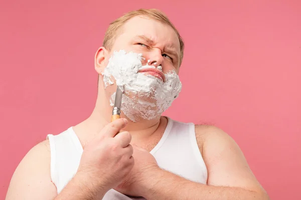 Plump man looks annoyed, feeling pain or irritation, while shaving with razor. — Stock Photo, Image