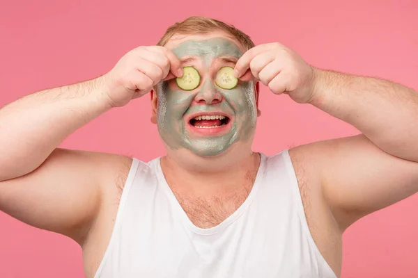 Hombre feliz con máscara de arcilla facial para el cuidado de la piel que se divierte con rodajas de pepino — Foto de Stock