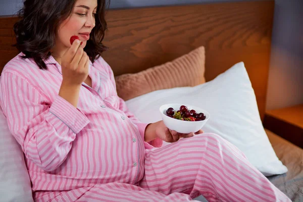 Wanita hamil makan stroberi duduk di tempat tidur di rumah — Stok Foto