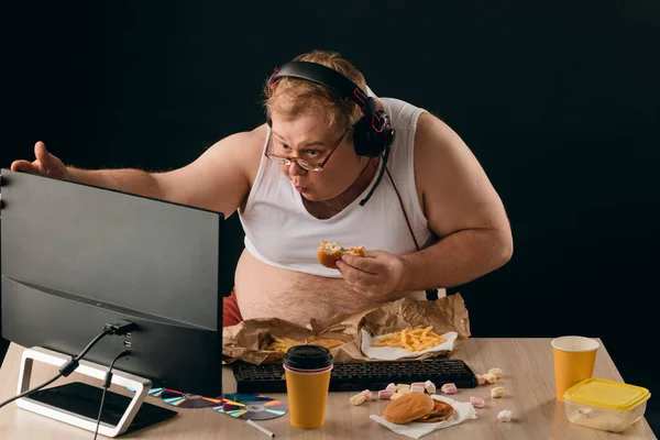 Engraçado homem gordo olhando para a tela do computador e comer um hambúrguer — Fotografia de Stock
