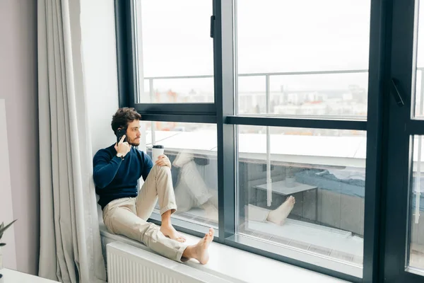 Schöner Mann sitzt mit Smartphone in der Hand auf der Fensterbank. — Stockfoto