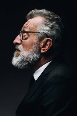 Yaşlı, düşünceli, klasik takım elbiseli bir işadamı gözlerini kaçırıp, karanlıkta poz veriyor.