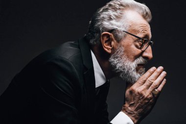 Yaşlı, düşünceli, klasik takım elbiseli bir işadamı gözlerini kaçırıp, karanlıkta poz veriyor.