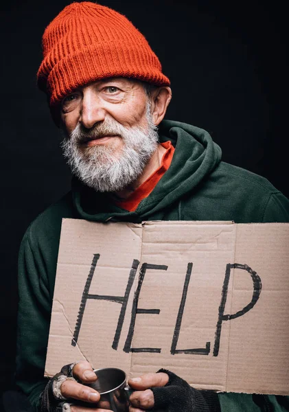 Triste vagabundo viejo agotado con sombrero rojo y ropa de calle sosteniendo un cartel de cartón. — Foto de Stock