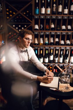 Şarap garsonu şarap şişelerine bakıyor ve şarap mahzeninde deftere yazıyor.
