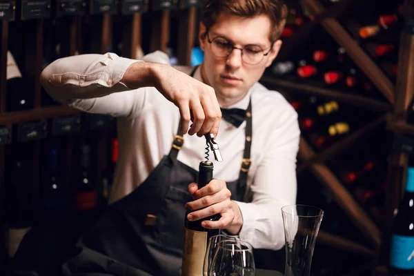 Bartender opening wine bottle with corkscrew in restaurant with wine shelves. — Stock Photo, Image