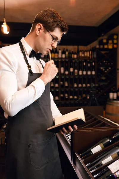 Şarap garsonu şarap şişelerine bakıyor ve şarap mahzeninde deftere yazıyor. — Stok fotoğraf