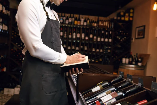 Male cavist examines the bottles with wine and making notes at his notepad