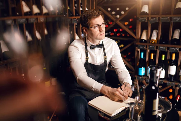 Επιστάτης κρασιού κοιτάζοντας μπουκάλια κρασιού και γράφοντας στο σημειωματάριο στο κελάρι κρασιού — Φωτογραφία Αρχείου