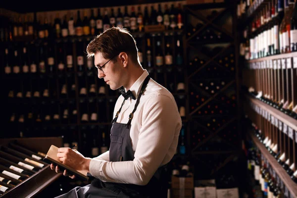 Ο φροντιστής αρσενικού οίνου εξετάζει φιάλες κρασιού και γράφει σε σημειωματάριο στην κάβα κρασιού — Φωτογραφία Αρχείου
