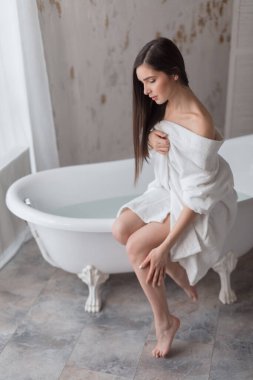 Genç kadın kısmen bornoz giymiş, banyo yaptıktan sonra bacaklarına bakıyor.