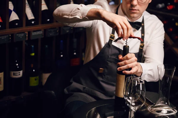 Bartender opening a wine bottle with corkscrew in restaurant with wine shelves. — Stock Photo, Image