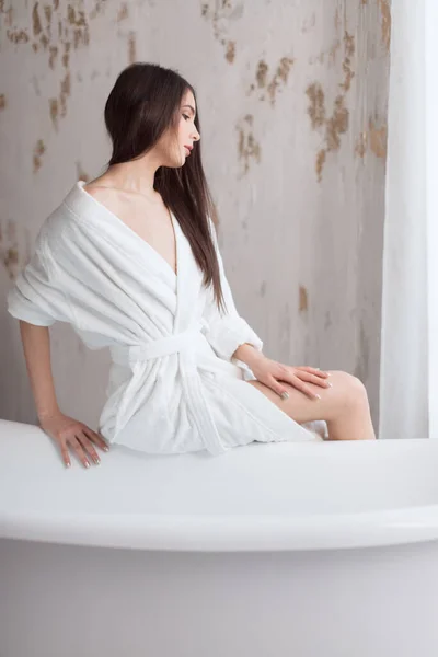 Gyönyörű fiatal hölgy fürdőköpenyben fog fürödni a fehér fürdőszobában. — Stock Fotó