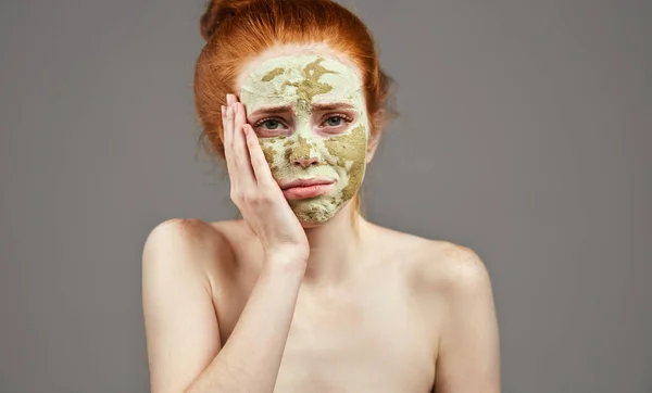 Δυστυχισμένη όμορφη νεαρή γυναίκα με πράσινη μάσκα προσώπου από πηλό που αγγίζει το μάγουλό της — Φωτογραφία Αρχείου