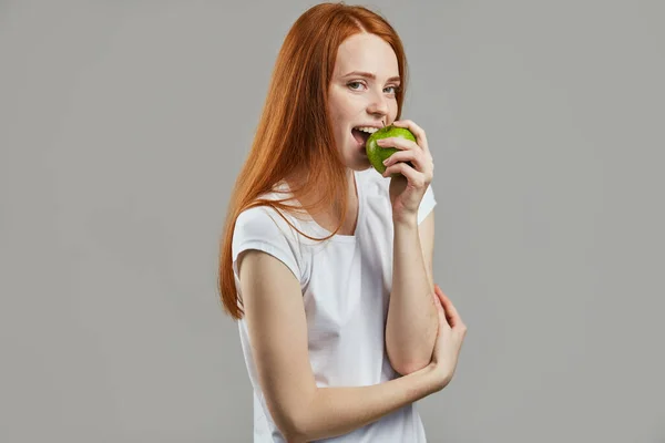 Impresionante chica con el pelo largo y rojo está comiendo una manzana verde —  Fotos de Stock