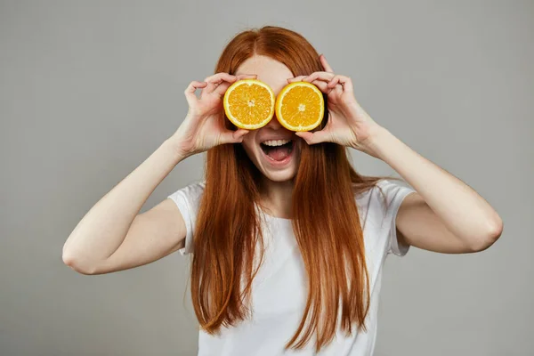 Feliz joven sosteniendo dos rebanadas de naranja en su cara sobre fondo gris — Foto de Stock