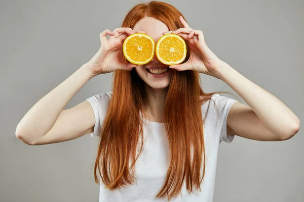 Agradable chica con el pelo largo y rojo sosteniendo naranjas cerca de la cara — Foto de Stock