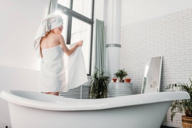 Kadın model, pencereyle küvet arasında duruyor. Çıplaklığını saklamak için havlu kullanıyor.