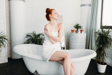 Kızıl saçlı kadın yeşil bitkilerle dolu geniş banyoda köpük banyosu hazırlıyor.