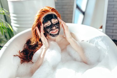 Yüzünde kilden bir maskeyle küvette yatan uzun kızıl saçlı bir kadın.