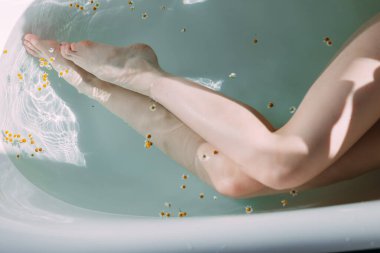 Küvette, temiz su ve küçük çiçeklerle kesilmiş ince kadın bacakları.