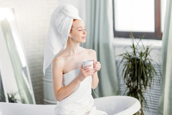 Glad kvinna i badrummet lyssnar på musik och dricker te sittande på badkar — Stockfoto