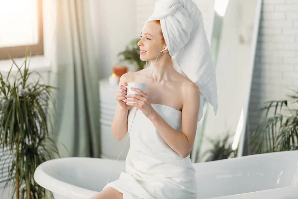 Szczęśliwa kobieta w łazience słucha muzyki i pije herbatę siedząc na wannie — Zdjęcie stockowe