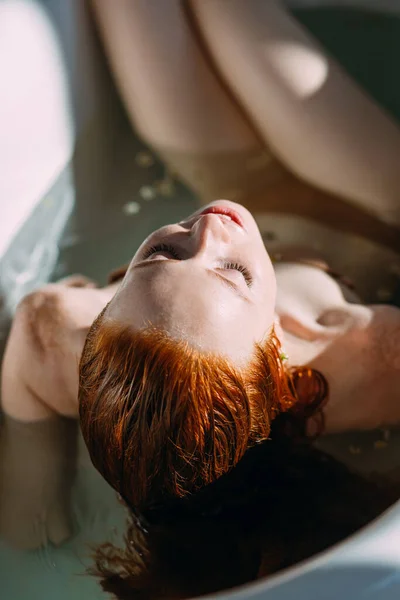 Mujer joven pelirroja acostada en la bañera, tocándose suavemente la cara con las manos — Foto de Stock