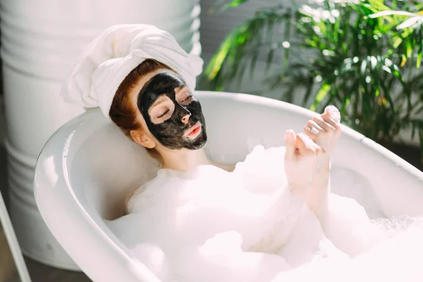 Vrouw met een handdoek op het hoofd liggend in bad met een kleimasker op haar gezicht — Stockfoto