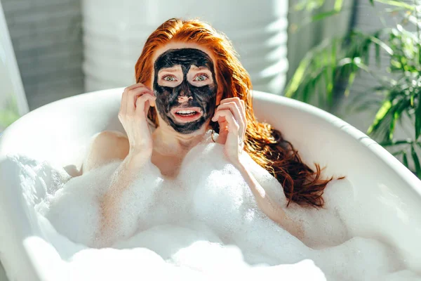 Žena s dlouhými zrzavými vlasy ležící ve vaně s hliněnou maskou na tváři — Stock fotografie