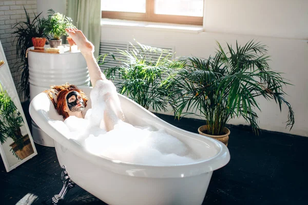 Donna con lunghi capelli rossi sdraiata nella vasca da bagno con una maschera di argilla sul viso — Foto Stock
