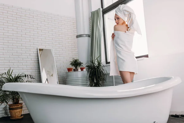 Mulher de cabelos vermelhos magro cobrindo seu corpo com toalha enquanto está de pé no banheiro — Fotografia de Stock