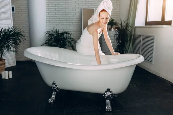 Mujer sonriente sentada en el borde de la bañera corriendo el baño, probando el agua con la mano —  Fotos de Stock