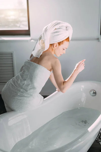 Glimlachende vrouw zitten op badkuip rand lopen het bad, het testen van water met de hand — Stockfoto