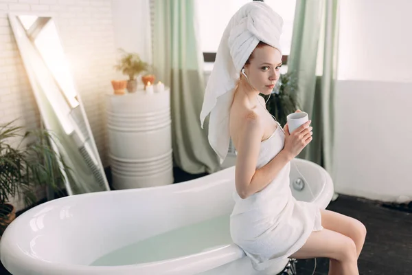Femme heureuse dans la salle de bain écouter de la musique et boire du thé assis sur la baignoire — Photo