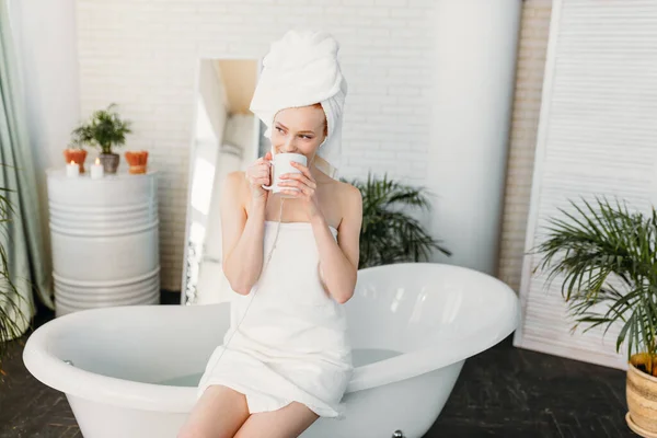 Femme heureuse dans la salle de bain écouter de la musique et boire du thé assis sur la baignoire — Photo