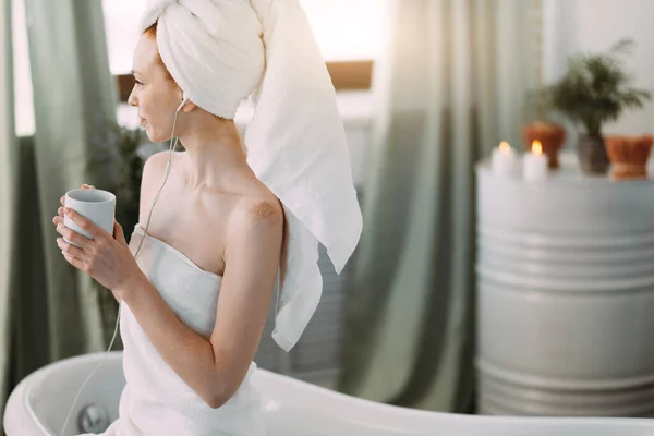 Щаслива жінка у ванній слухає музику і п'є чай, сидячи на ванні — стокове фото